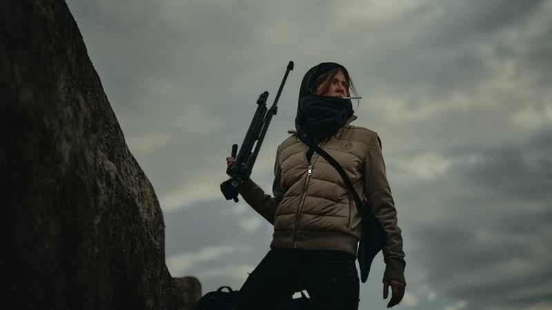 A woman with a scarf over her face carrying a rifle on a cliff path under storm cloud