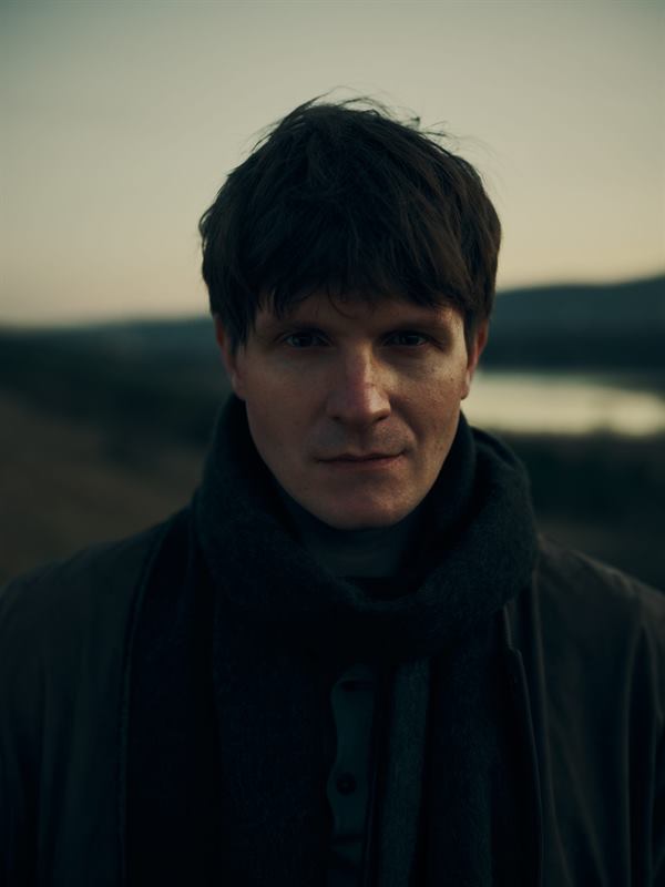 A man in a dark scarf standing before a hazy valley at dusk