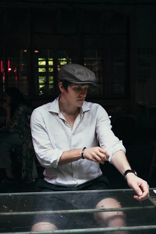 A person in a flat cap and white shirt leaning on a table outside a lit bar