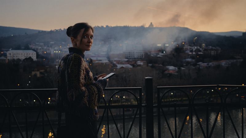 A woman at a railing above a smoke-covered city at sunset