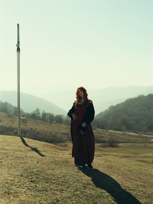 A woman in a long red dress on a hillside beside a tall pole