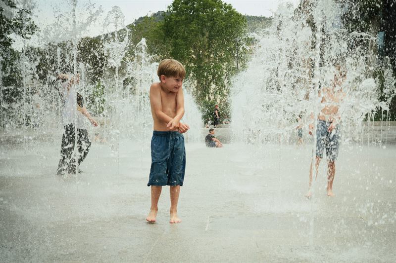 A boy standing in the spray of a fountain while other children run past
