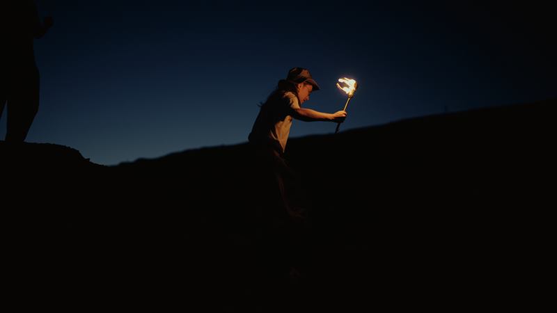A woman in a cap climbing a dark ridge at blue hour, a burning torch held out ahead of her