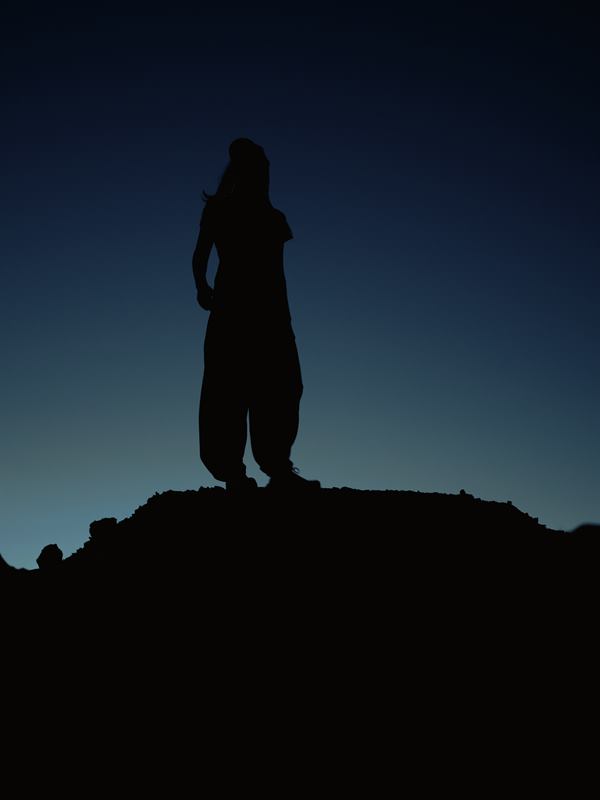 A woman standing alone on a ridge, in full silhouette against the last teal light of dusk