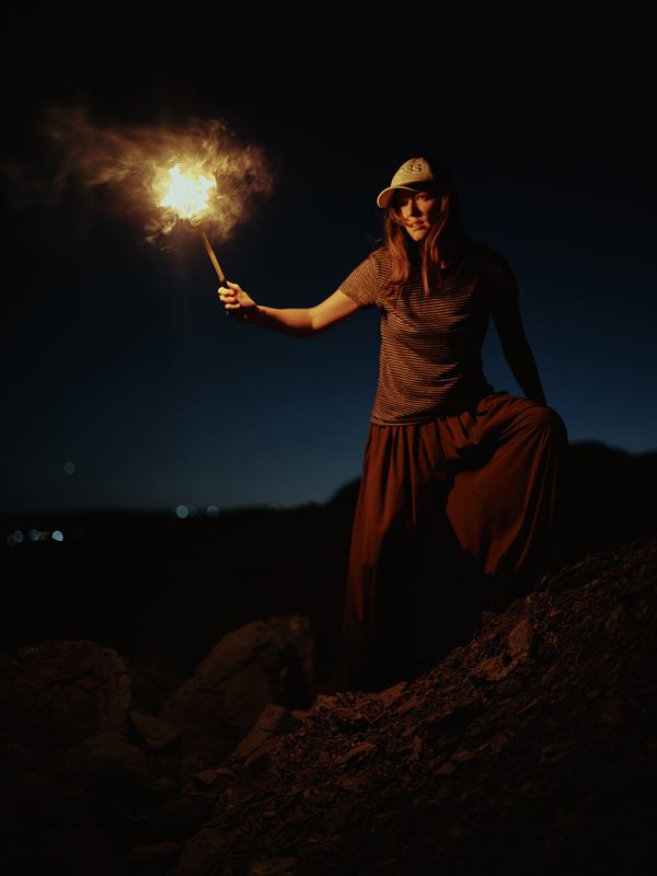 A woman holding a burning torch on a rocky slope, the flame lighting her face, city lights far below