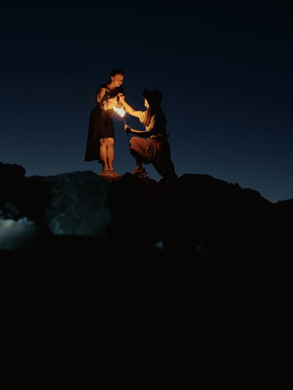 Two people on a dark ridge at night, one kneeling to pass a lit torch to the other