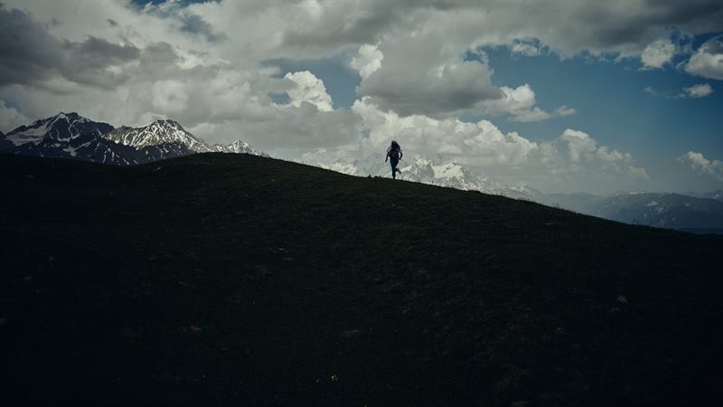 A lone walker on a dark ridge, snowy peaks and heavy cloud behind