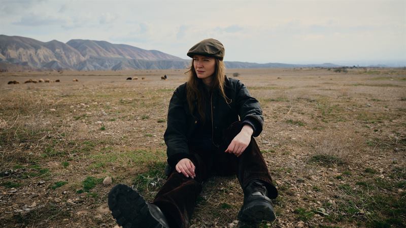A woman in a flat cap sitting on dry grassland, hills behind her