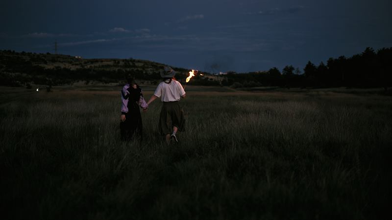 A couple walking hand in hand through tall grass at dusk, a lit torch carried ahead of them