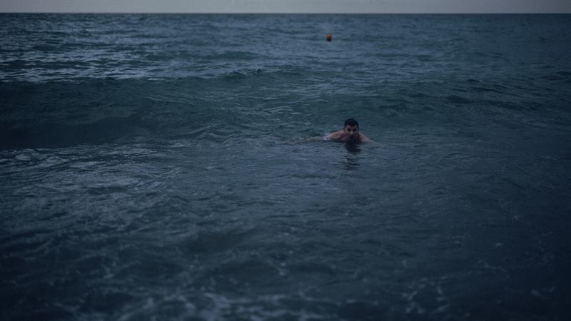 A lone swimmer far out in an open grey sea at dusk, a marker buoy on the horizon