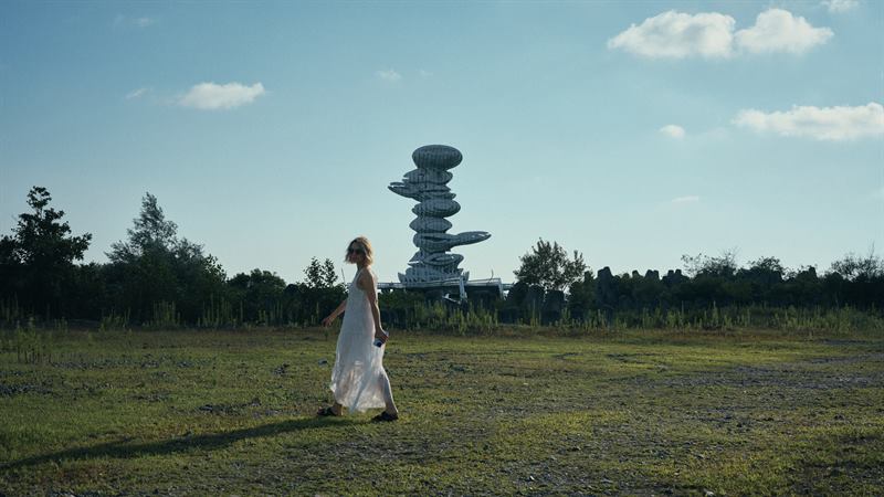 A woman in a white dress crossing a summer field below the spiral tower at Anaklia, Georgia