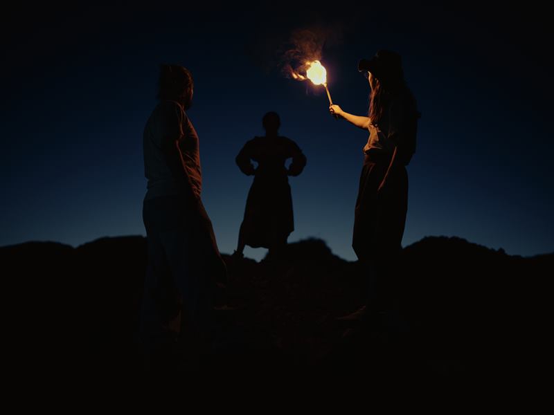 Three people on a hilltop at blue hour, one of them raising a burning torch between them
