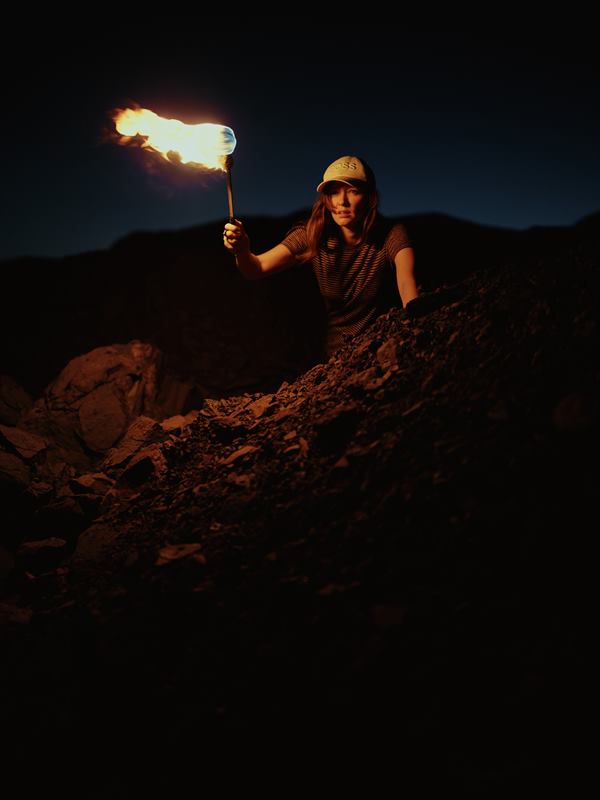 A woman raising a burning torch above a slope of broken rock at nightfall