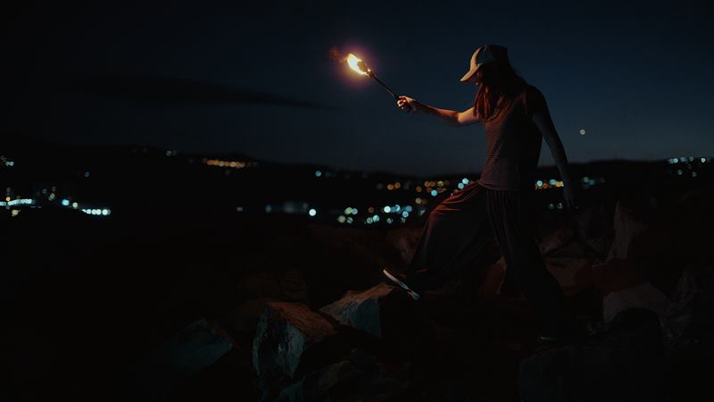 A woman stepping across boulders with a lit torch, city lights spread out behind her