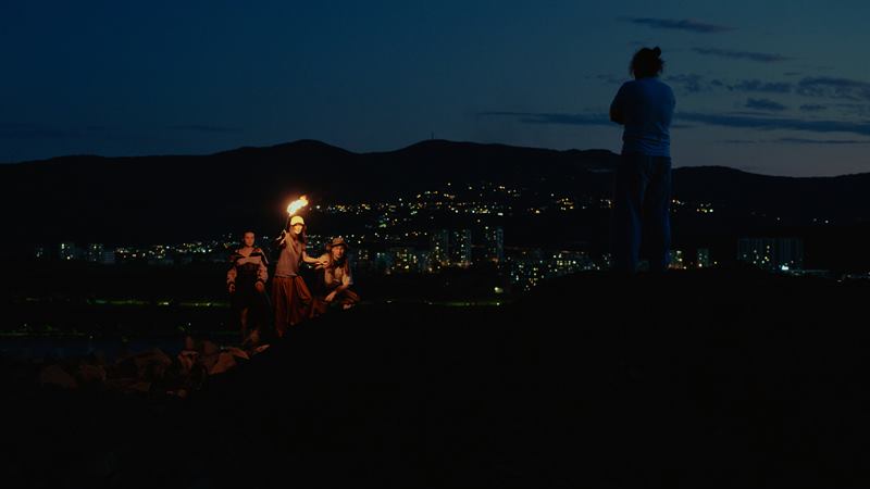 A group gathered around a torch on a hill above a city at blue hour, one figure standing apart