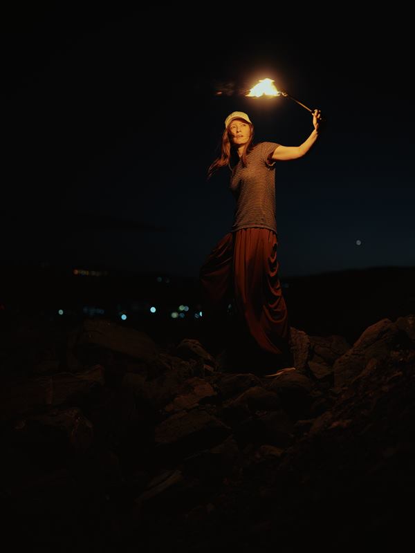A woman holding a torch overhead on a rocky ridge, city lights scattered in the dark below
